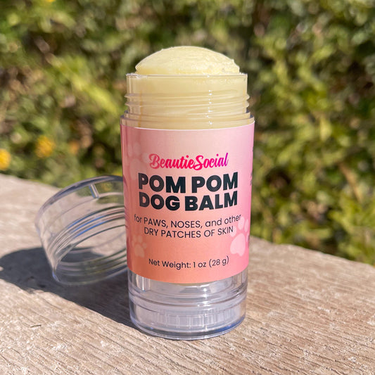 A 1 oz clear plastic tube of BeautieSocial Pom Pom Dog Balm with a pink label, sitting outside with a blurred green garden background. Tube is open with cap sitting beside it.