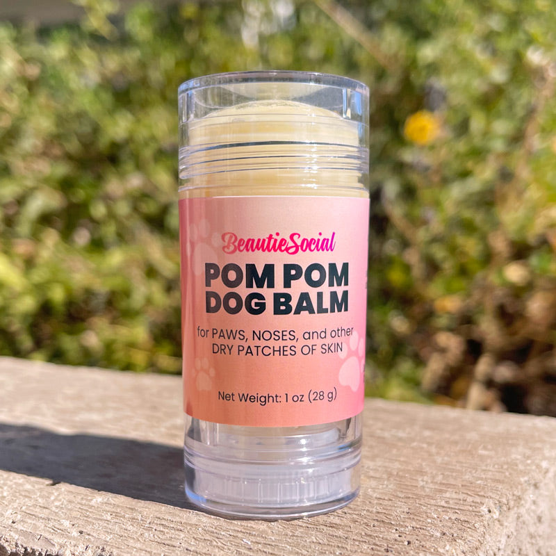 A 1 oz clear plastic tube of BeautieSocial Pom Pom Dog Balm with a pink label, sitting outside with a blurred green garden background.