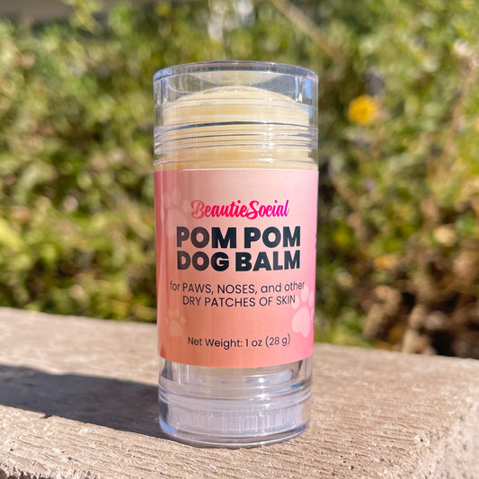 A 1 oz clear plastic tube of BeautieSocial Pom Pom Dog Balm with a pink label, sitting outside with a blurred green garden background.