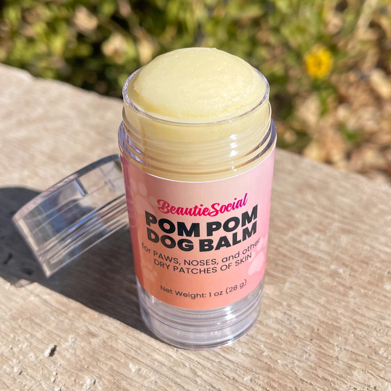 Looking down at a 1 oz clear plastic tube of BeautieSocial Pom Pom Dog Balm with a pink label, sitting outside with a blurred green garden background. Tube is open with cap sitting beside it.