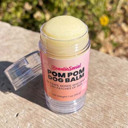 Looking down at a 1 oz clear plastic tube of BeautieSocial Pom Pom Dog Balm with a pink label, sitting outside with a blurred green garden background. Tube is open with cap sitting beside it.