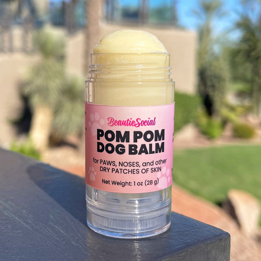 A clear plastic open tube of BeautieSocial Pom Pom Dog Balm with a pink label, sitting on a dark stone surface with a blurred outdoor garden background.