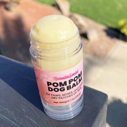 A clear plastic open tube of BeautieSocial Pom Pom Dog Balm with a pink label, filled with pale yellow paw balm, sitting on a dark surface outdoors.